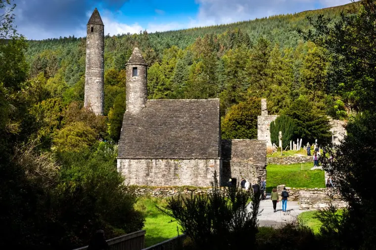 Glendalough round tower and the stone roofed St Kevin's Church below a forested hillside, with visitors on the paths