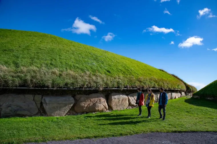 Three visitors walking beside the grass covered mound and large kerbstones of Knowth under a blue sky